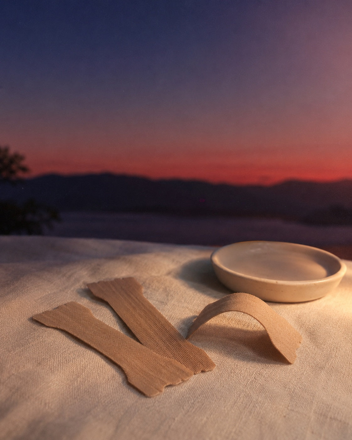 Three unbranded nude-tan fabric nasal strips fanned on ivory linen at dusk, a small cream ceramic dish beside them.
